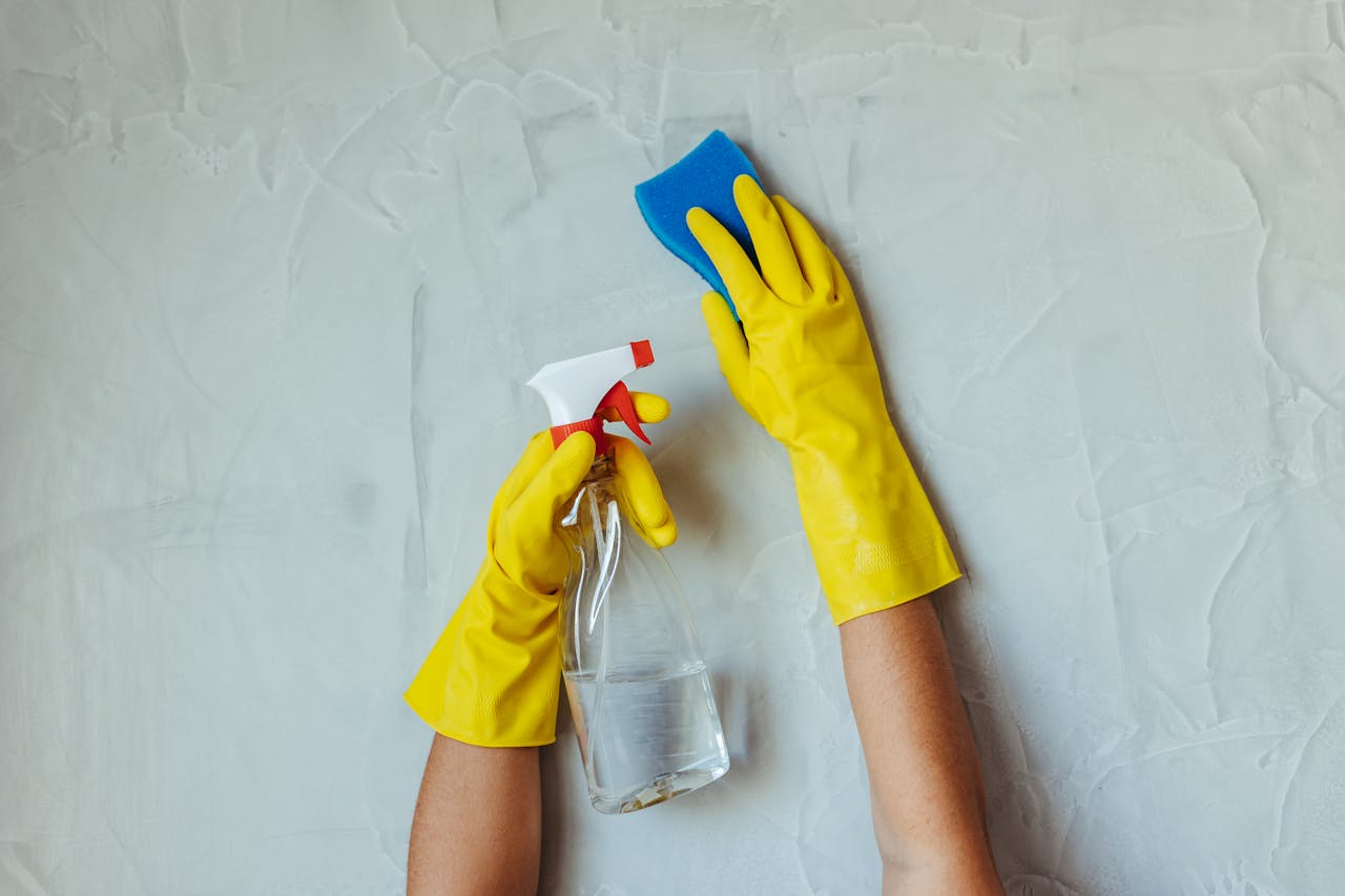hero-img-01 Close-up of hands in yellow gloves cleaning a wall with a spray bottle and sponge.