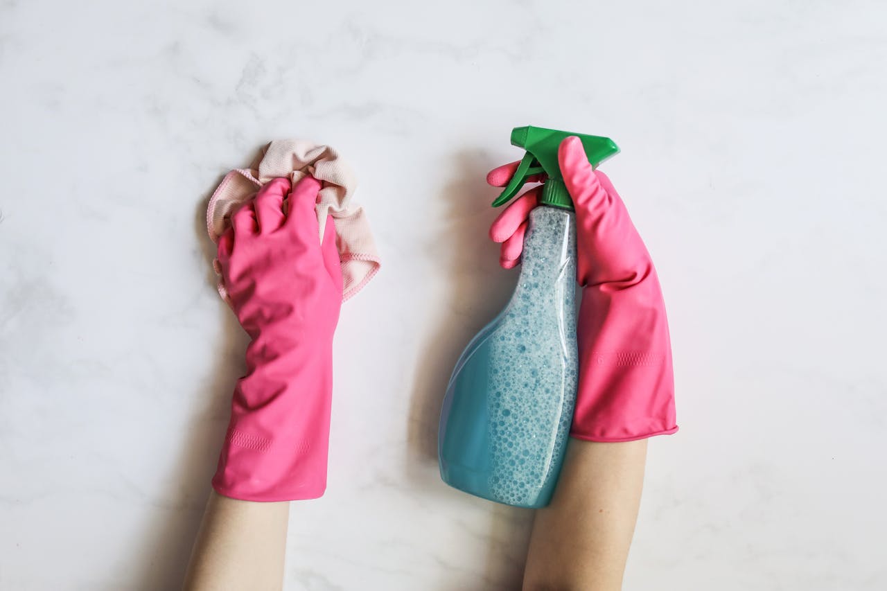 hero-img-02 A pair of hands in pink gloves are cleaning with a spray bottle on a white background.